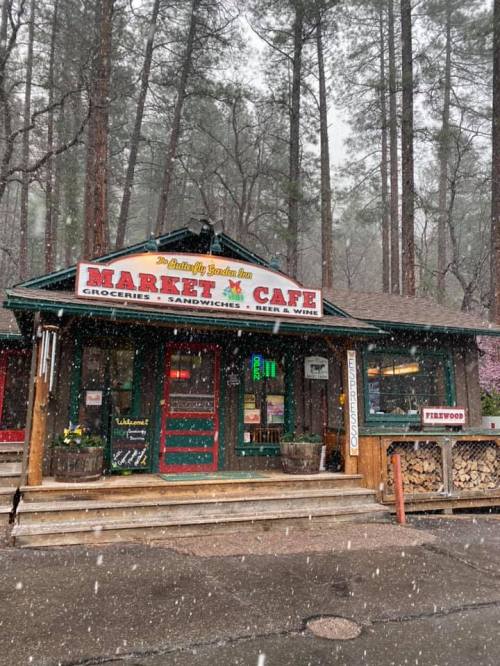 A cozy market café nestled in the woods, with snow falling gently around it. Firewood stacked outside.