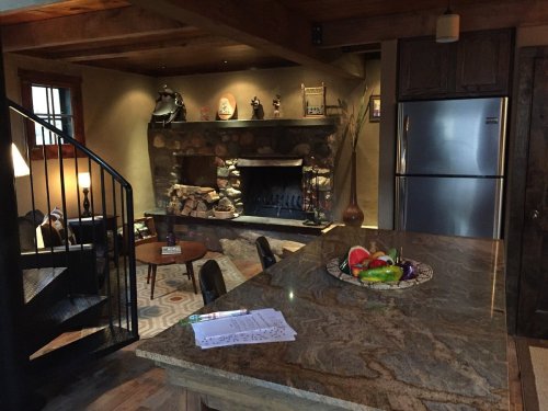 Cozy living space with a stone fireplace, wooden beams, and a kitchen island featuring a fruit bowl.