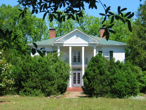 A white two-story house with a green roof, surrounded by lush greenery and trees on a sunny day.