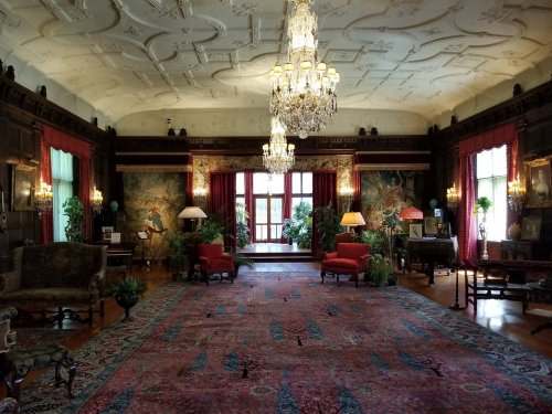 Luxurious room with ornate ceiling, chandeliers, red curtains, vintage furniture, and lush plants. Richly patterned carpet.