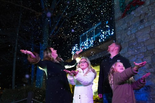 A family joyfully catches falling snowflakes, surrounded by festive lights in a winter setting.