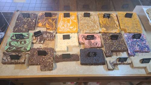 A display of various colorful fudge flavors, each labeled with a small black sign, arranged on a wooden counter.