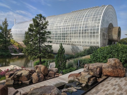 A modern glass greenhouse surrounded by greenery and rocks, reflecting a serene landscape under a blue sky.