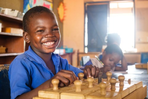 A smiling boy in a blue shirt plays chess at a table, with another child focused in the background.