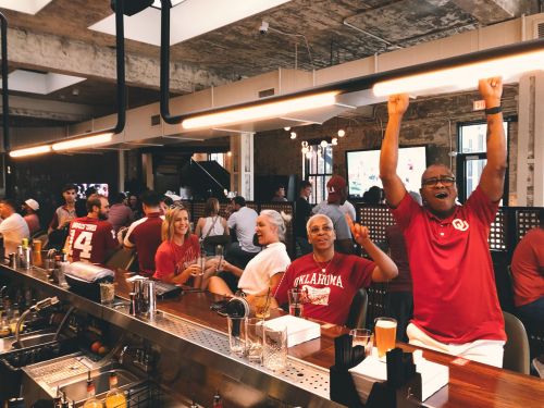 A lively bar scene with patrons celebrating, wearing red, and enjoying drinks while watching a game on TV.
