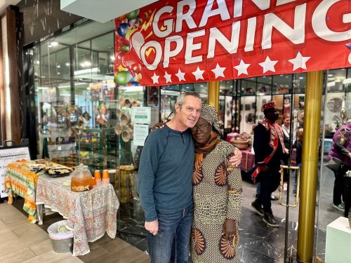 A man and a woman in traditional attire pose together in front of a "Grand Opening" banner at a festive event.