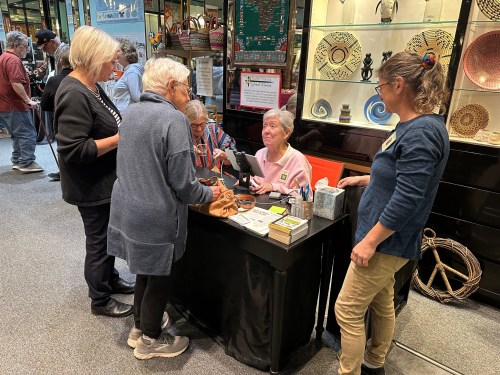 A group of people at a vendor table, engaging with a staff member in a busy market setting.