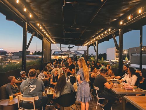 A lively outdoor dining area filled with people enjoying food and drinks under string lights at sunset.