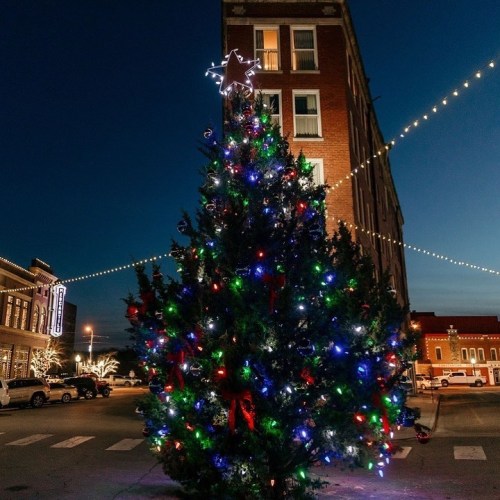 A brightly lit Christmas tree with decorations stands in front of a historic building at dusk, with festive lights above.