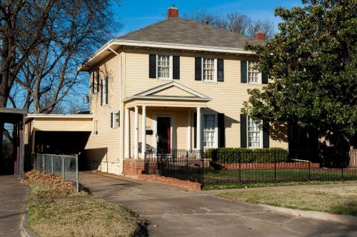 A two-story yellow house with black shutters, a front porch, and a carport, surrounded by trees and a lawn.
