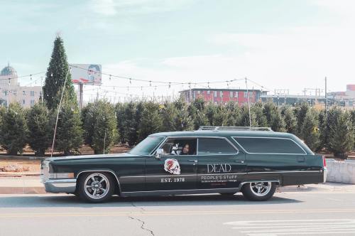 A vintage hearse with "Dead People's Stuff" written on the side, parked on a street lined with trees.