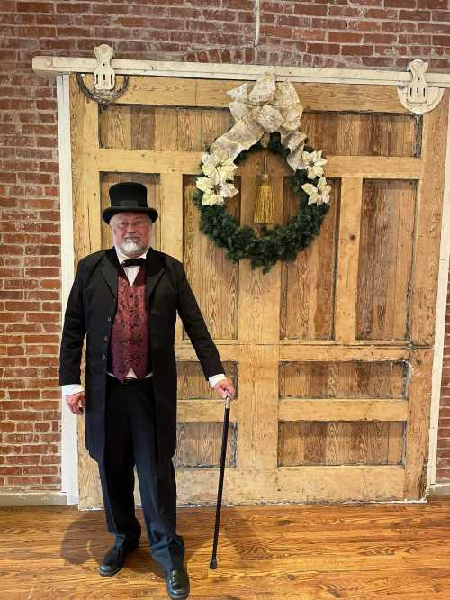 A man in a formal black coat and top hat stands by a rustic wooden door adorned with a festive wreath.