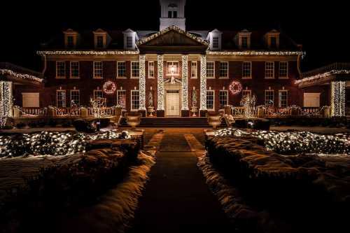 A beautifully decorated brick building illuminated with festive lights and wreaths, surrounded by snowy landscaping at night.