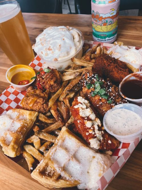 A wooden platter filled with fried chicken, waffles, fries, sauces, and a dessert, alongside a beer and a colorful drink.
