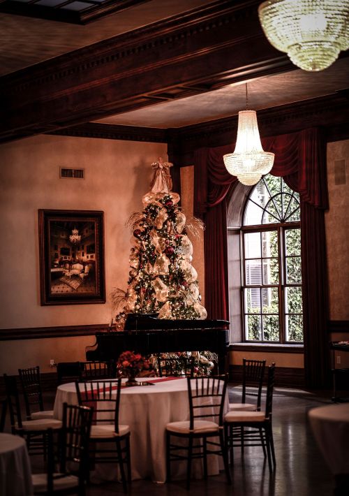 Elegant indoor setting with a decorated Christmas tree beside a grand piano, surrounded by tables and soft lighting.