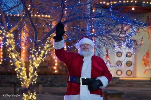 Santa Claus waves cheerfully in front of a tree adorned with blue and white lights during the holiday season.