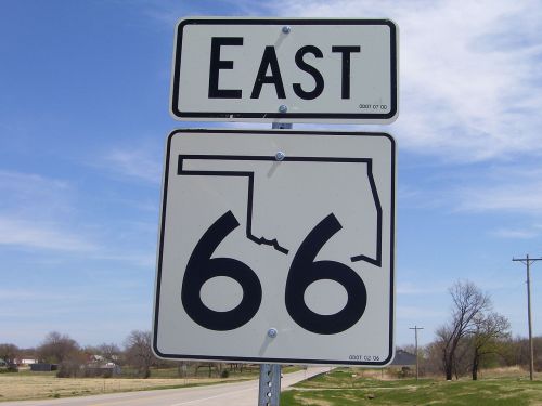 Sign indicating Route 66 East in Oklahoma, featuring a stylized outline of the state. Clear blue sky in the background.