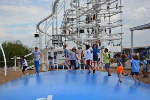 Children jumping on a large blue inflatable surface, with a climbing structure and people in the background.