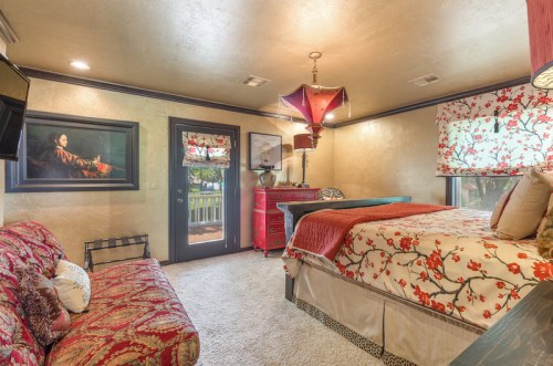 Cozy bedroom featuring a bed with floral bedding, a red accent chair, and decorative lamps, with a painting on the wall.