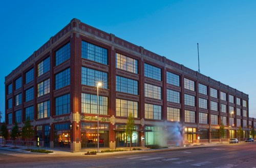A large brick building with many windows, illuminated at dusk, featuring a restaurant on the ground floor.