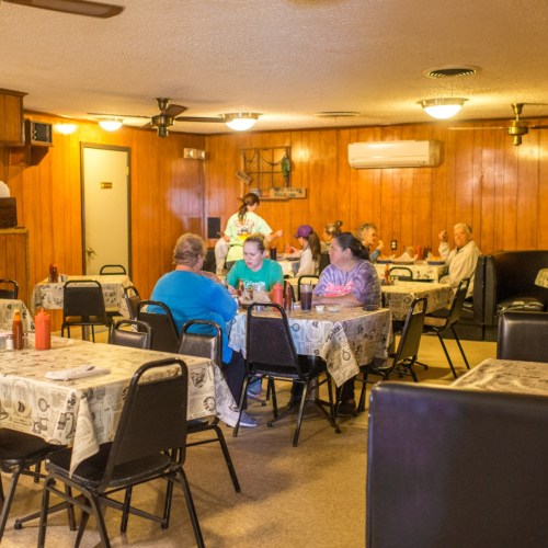 A cozy diner interior with several tables, patrons seated, and a staff member serving food. Warm lighting and wooden decor.