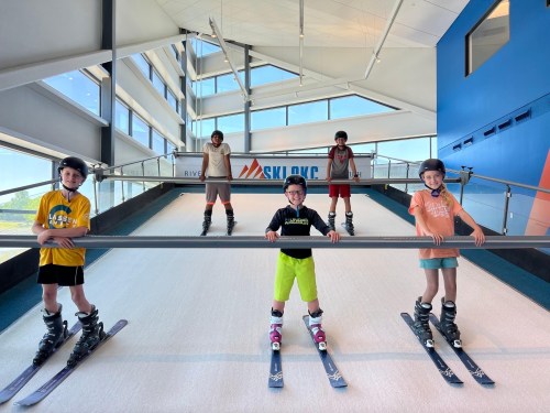 Five children in ski gear stand on a ski slope indoors, smiling and posing with a railing in a modern facility.