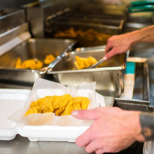 A hand holds a takeout container with fried food, while trays of similar food are visible in the background.