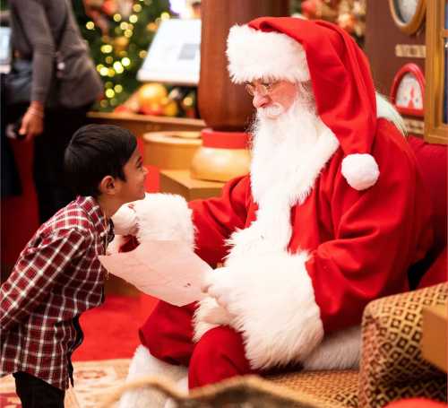 A child in a plaid shirt smiles at Santa Claus, who is dressed in a red suit and reading a letter.