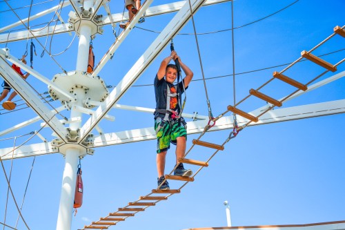 A child balances on a rope bridge in an outdoor adventure park, smiling against a clear blue sky.
