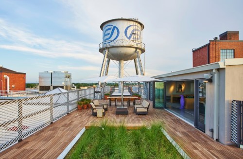 Rooftop terrace with seating area, greenery, and a water tower in the background under a clear sky.