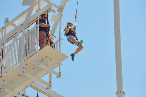 A person in a harness swings from a platform, with an operator overseeing from above on a sunny day.