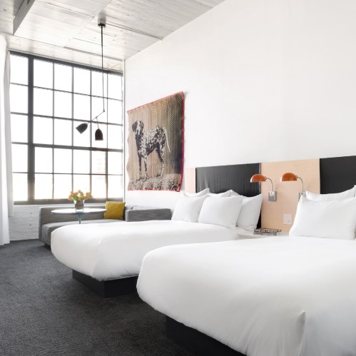 A modern hotel room with two beds, large windows, and stylish decor featuring a dog painting and cozy seating area.