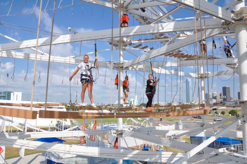 A group of people navigate a high ropes course, suspended above the ground, with a city skyline in the background.