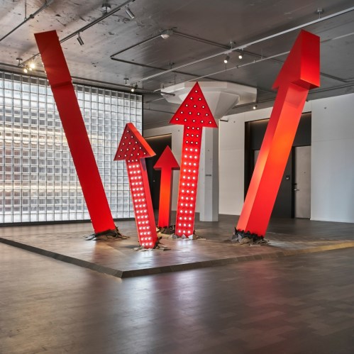 Red arrow sculptures of varying heights, illuminated with lights, in a modern gallery space with a minimalist design.