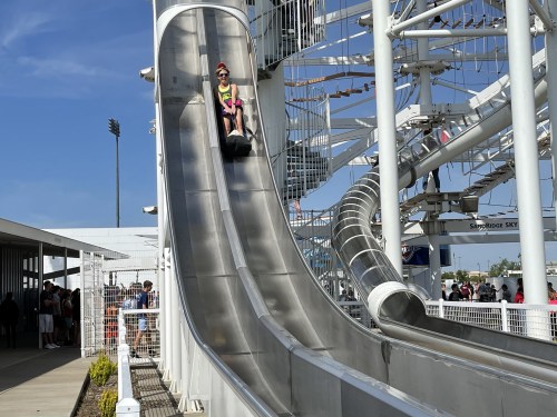 A person slides down a metal slide at an amusement park on a sunny day, with a crowd in the background.