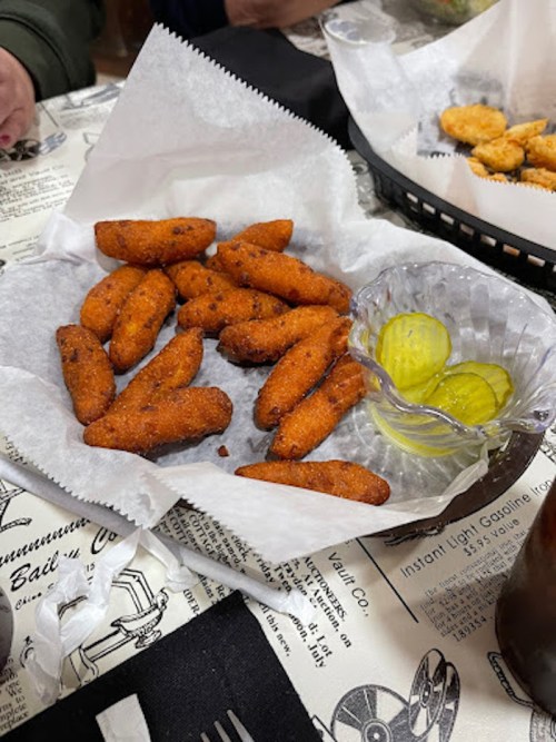 A basket of golden-brown fried snacks with a side of pickles on a paper-lined plate.
