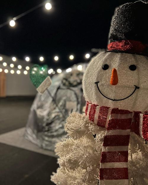 A cheerful snowman with a scarf and hat stands in front of a decorated area with string lights and a heart-shaped ornament.