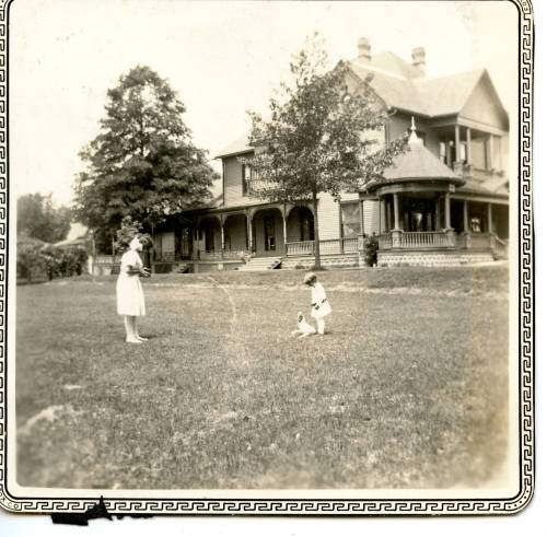 A woman and a child play in a grassy yard in front of a large, historic house. Trees surround the area.