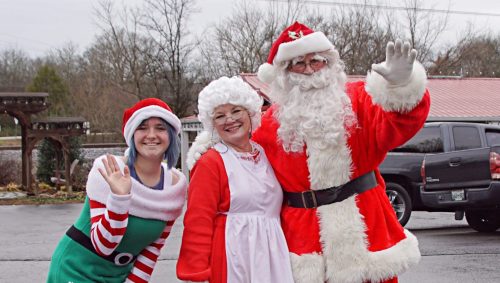 A woman in a Santa hat and elf costume poses with Santa and Mrs. Claus, all smiling and waving outdoors.