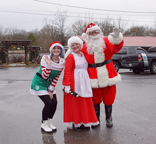 A cheerful Santa, Mrs. Claus, and an elf pose together, waving in a festive outdoor setting.