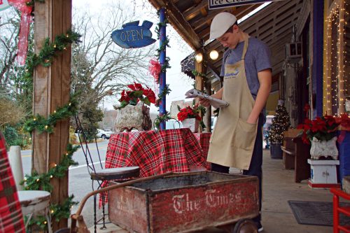 A young man in an apron stands by a festive outdoor ice cream shop, looking at a cart decorated for the holidays.
