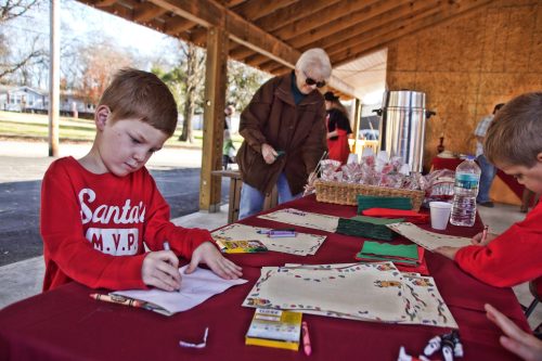 A child in a Santa shirt colors at a table, while an older woman looks on in a festive outdoor setting.