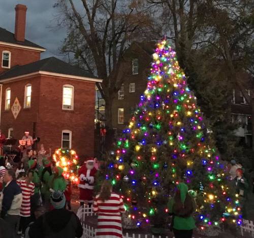 A festive scene with a brightly lit Christmas tree, Santa, and people in holiday attire celebrating in the evening.