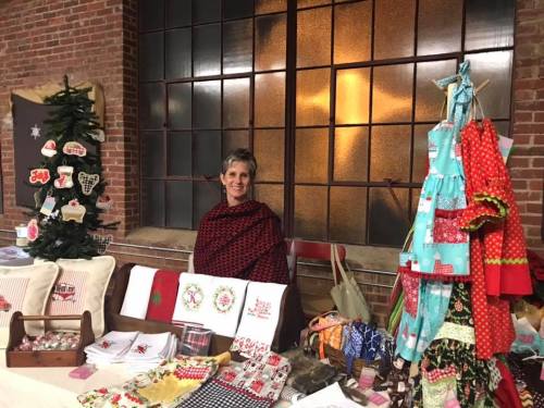 A woman in a red shawl stands behind a table filled with handmade crafts and decorations in a cozy setting.