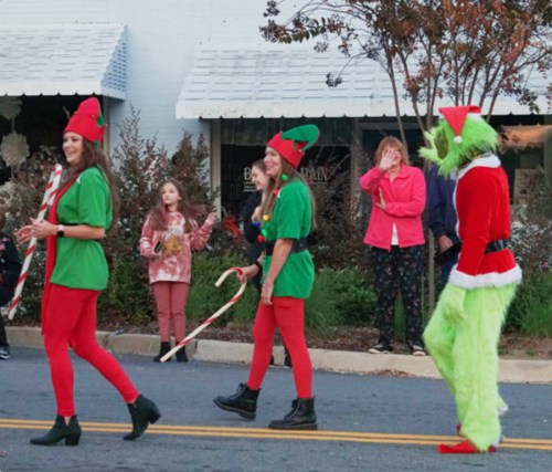 A festive scene with people in elf costumes and the Grinch, celebrating in a holiday parade.
