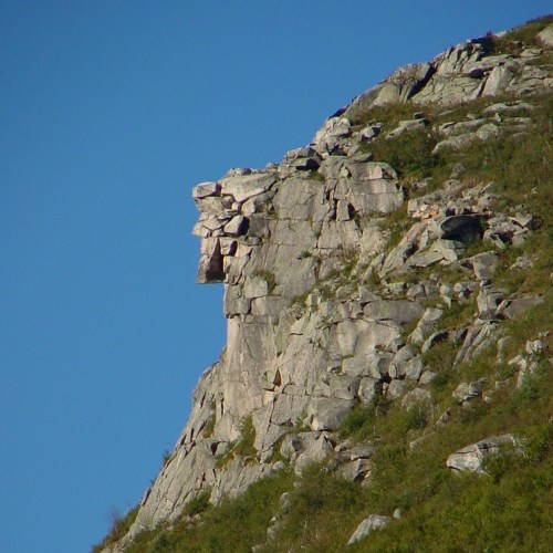 A rocky cliff formation resembling a profile of a face against a clear blue sky.