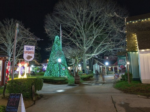 A festive night scene featuring a lit Christmas tree and holiday decorations in a charming outdoor plaza.