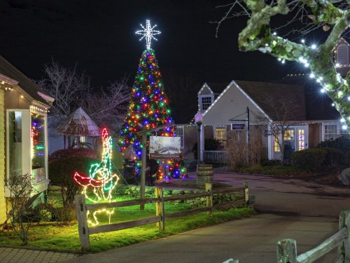 A brightly lit Christmas tree with colorful lights stands in a festive village at night, surrounded by holiday decorations.