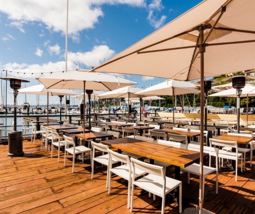 Outdoor dining area with wooden tables and umbrellas, overlooking a marina with boats under a blue sky.