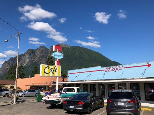 A colorful café sign with mountains in the background, featuring parked cars and a clear blue sky.